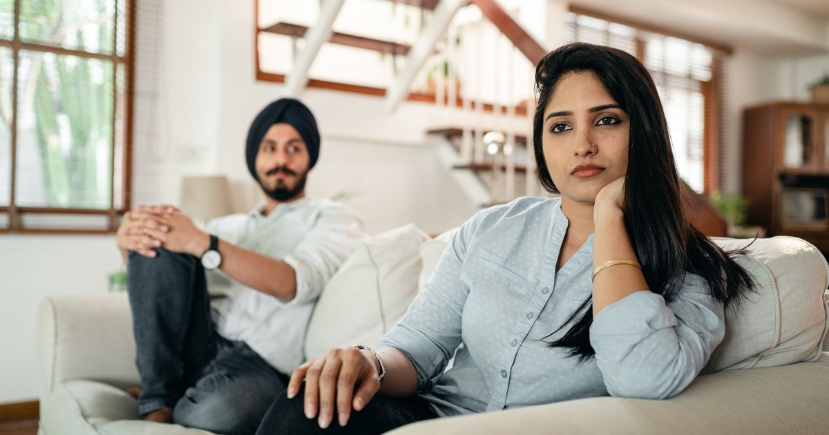 Offended Indian woman sitting on sofa with husband on another end and looking away while having argument