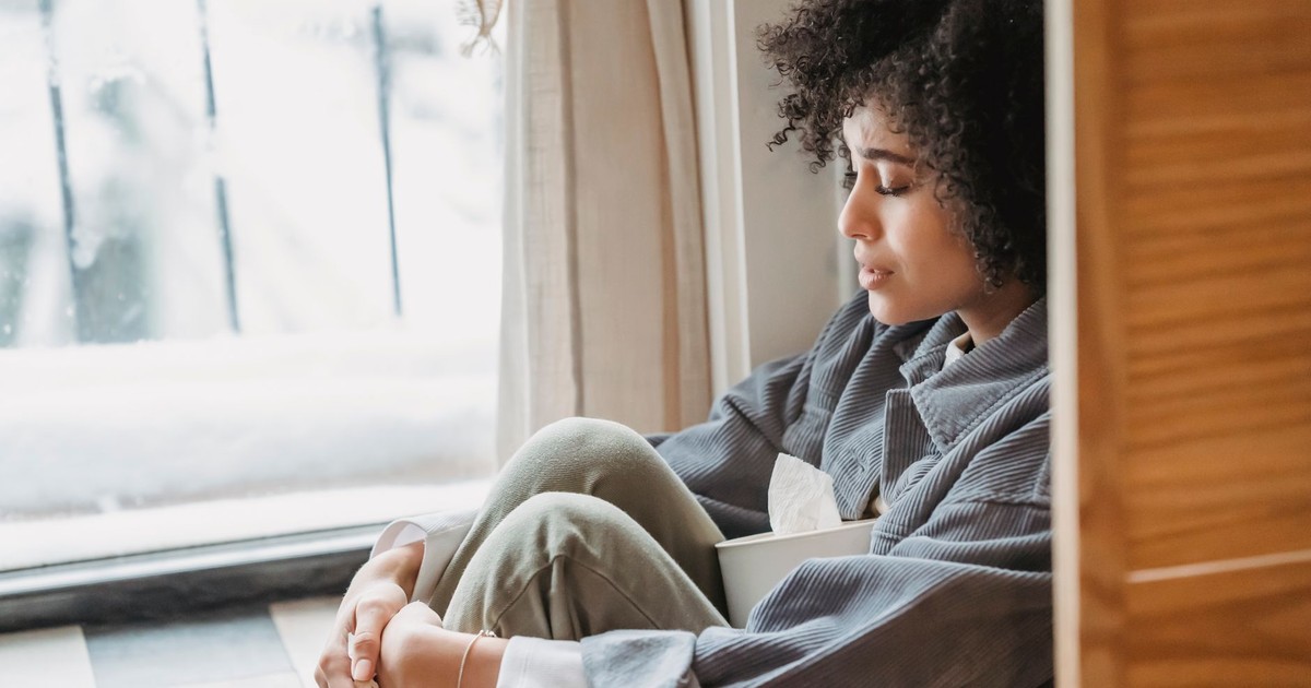 High angle side view of crying African American female with curly hair sitting on windowsill with box of tissues
