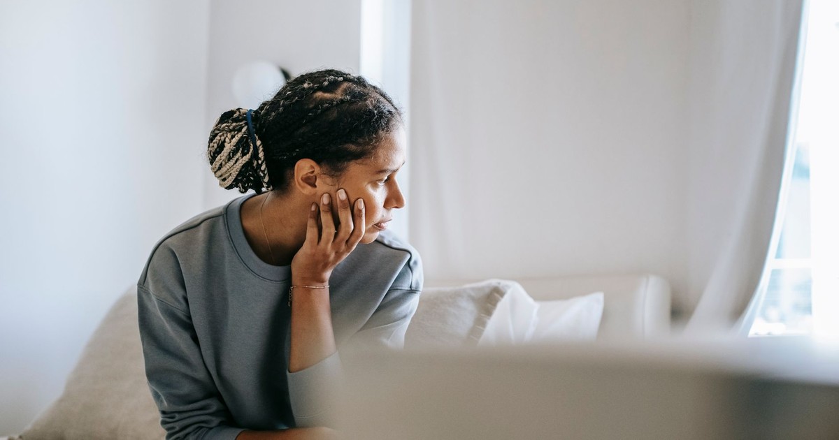 Young black female in casual outfit sitting on cozy white bed and thinking while looking away in daytime