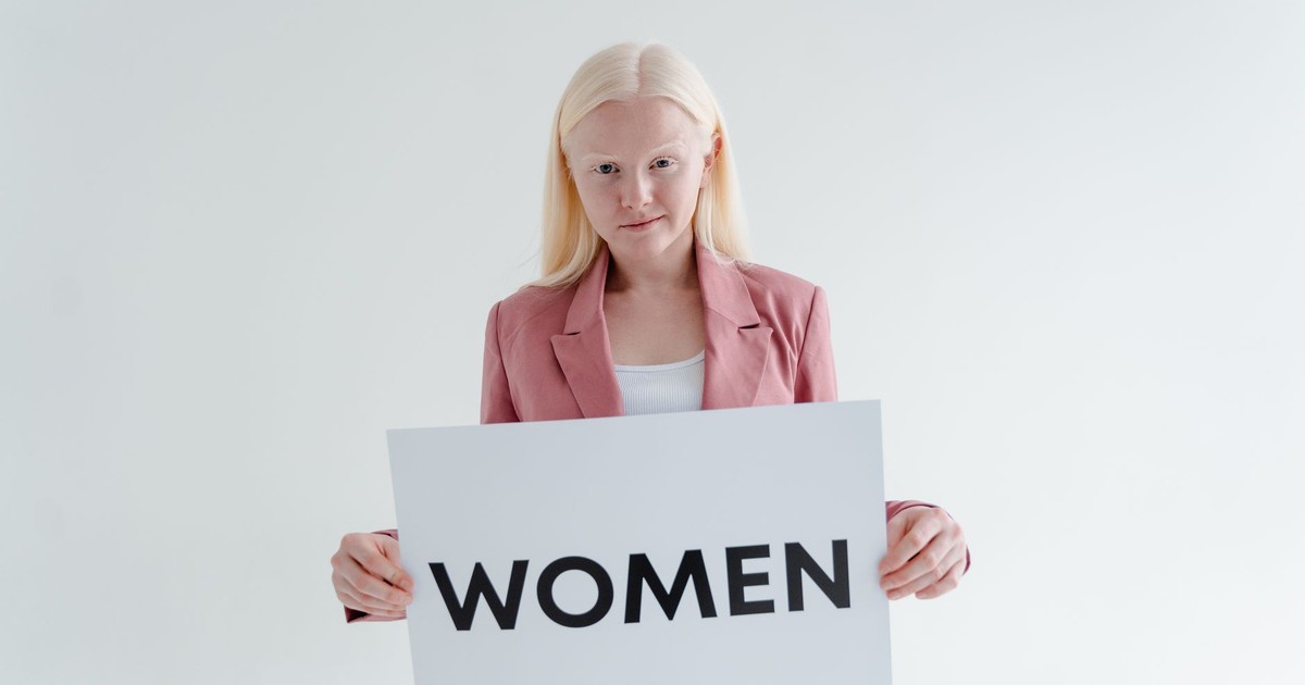Confident woman in pink blazer holds a sign reading 'Women' on a white background.
