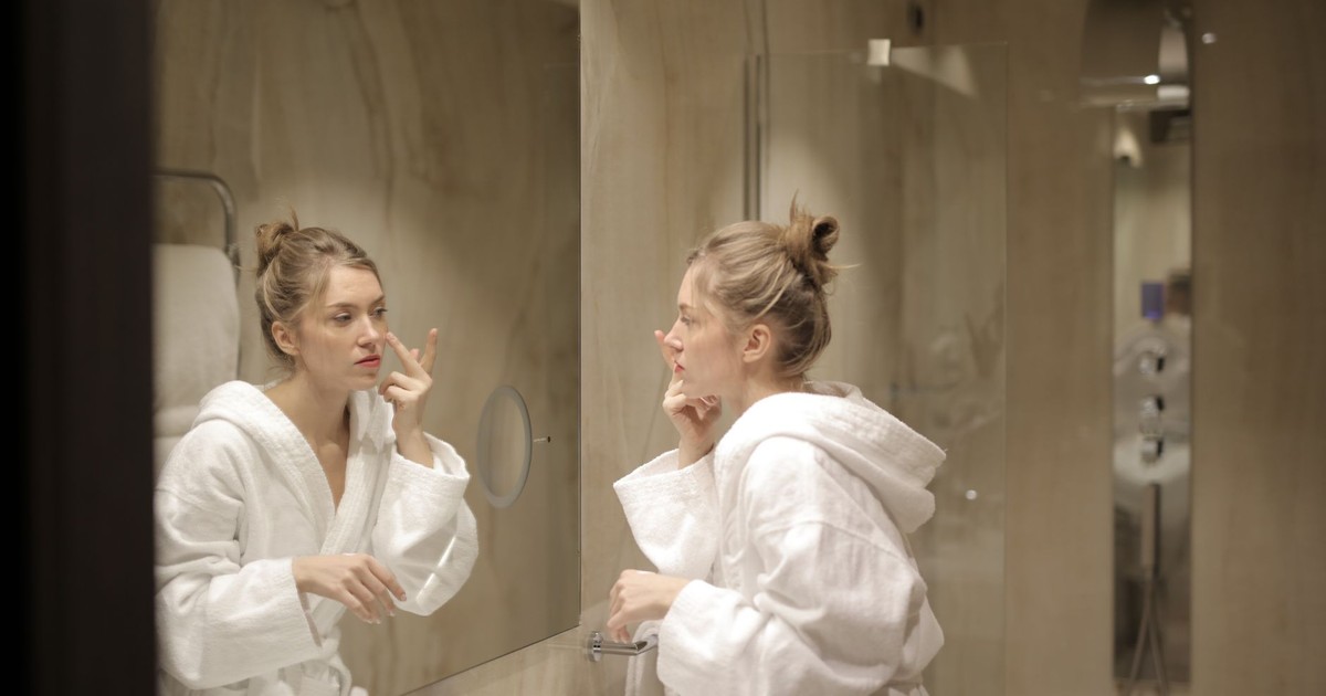 A woman in a white bathrobe applies moisturizer while looking at her reflection in the bathroom mirror.