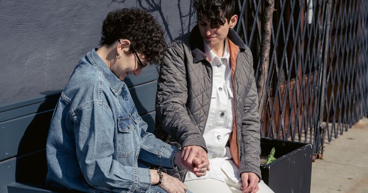 Positive young lesbian couple with short dark hair in casual outfits holding hands and talking with each other while resting on bench on city street