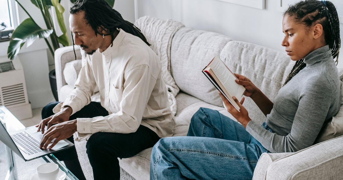 Side view of young black woman with Afro braids in casual outfit reading interesting novel while relaxing on comfortable couch near concentrated husband working remotely on laptop