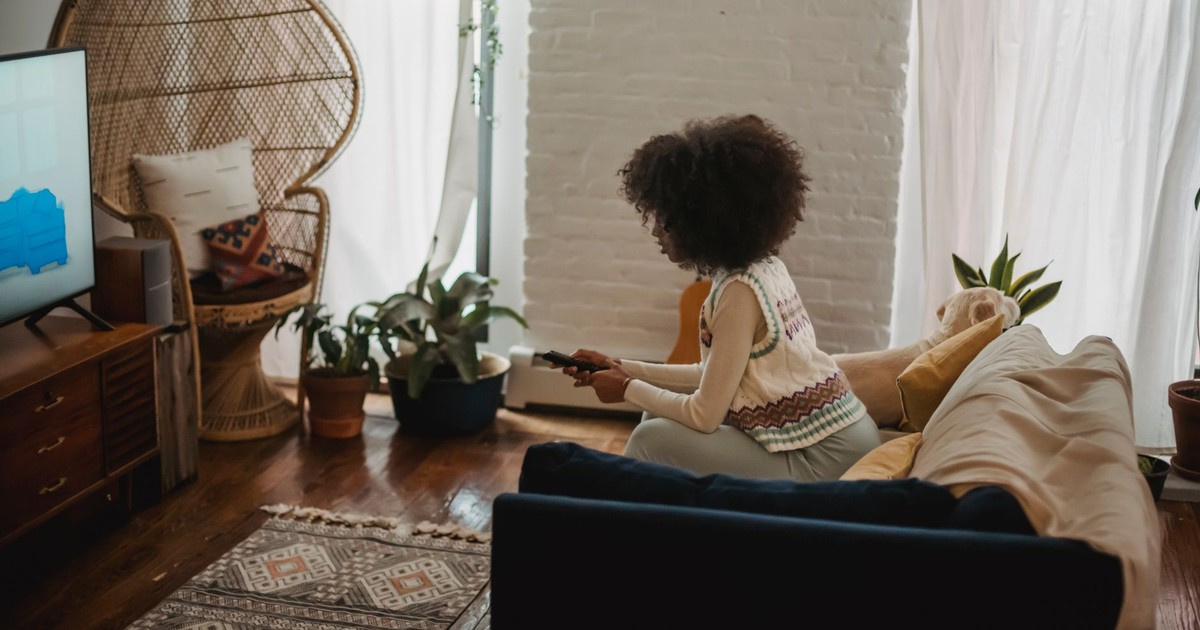 Side view of young African American female millennial with dark curly hair in stylish clothes switching channels on TV with remote controller while resting on sofa during weekend at home