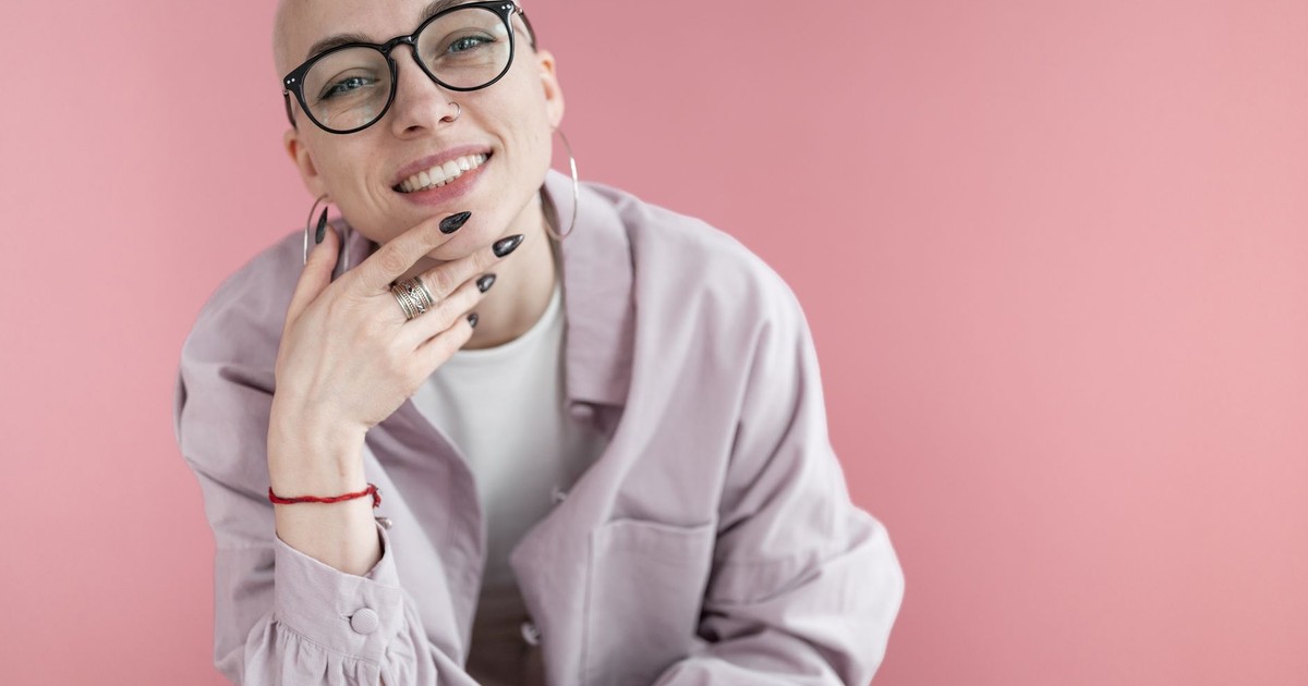 Smiling woman with short blond hair in trendy clothes and eyeglasses touching chin and looking at camera against purple background in studio