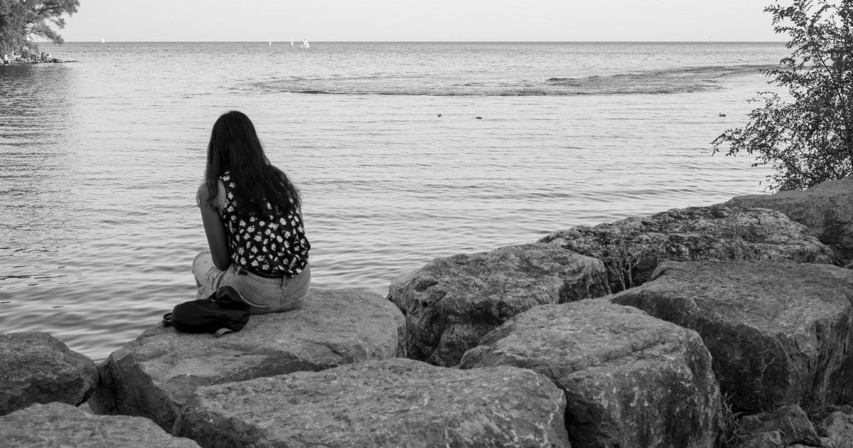 A woman sits on rocks gazing at the calm sea, capturing a serene moment of solitude and reflection.