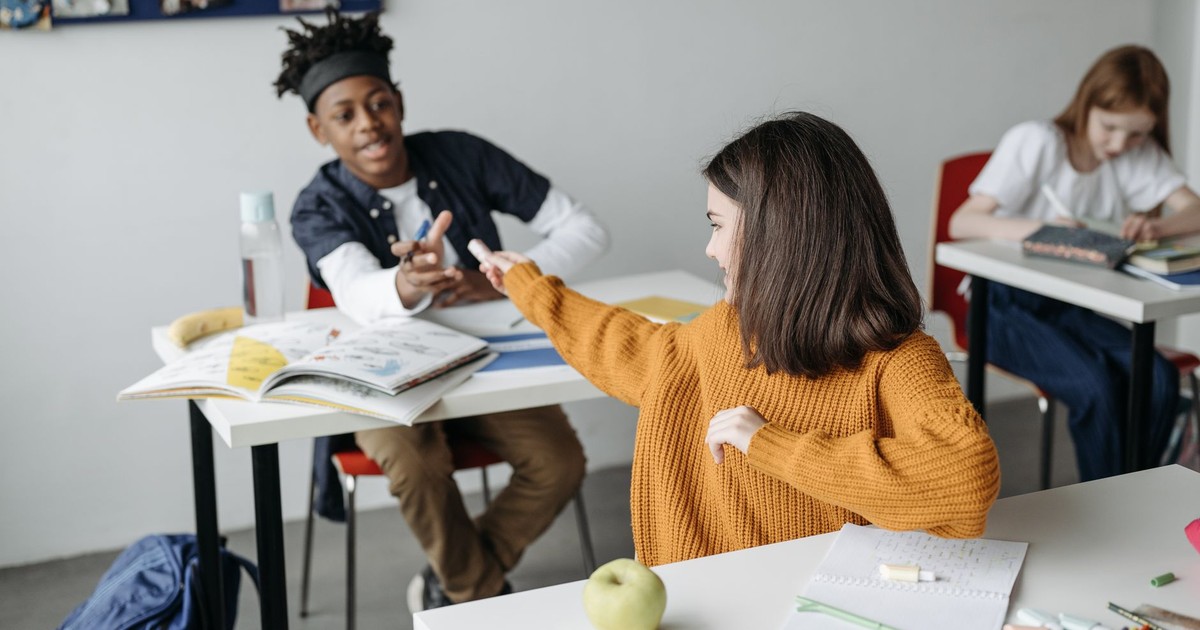 Students interacting in a classroom with textbooks and school supplies on desks.