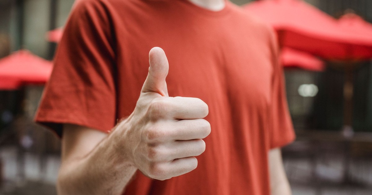 Crop anonymous young male in red t shirt showing thumb up gesture while standing on city street on sunny day