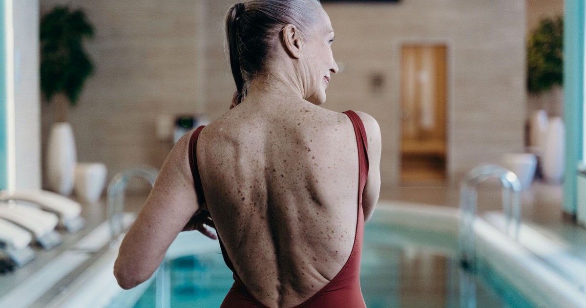Senior woman in red swimsuit standing at indoor pool side, exuding confidence.