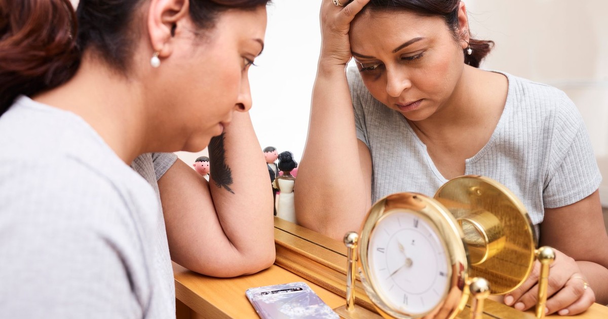 A woman looks pensive and tired, reflecting on burnout with a clock and mirror.
