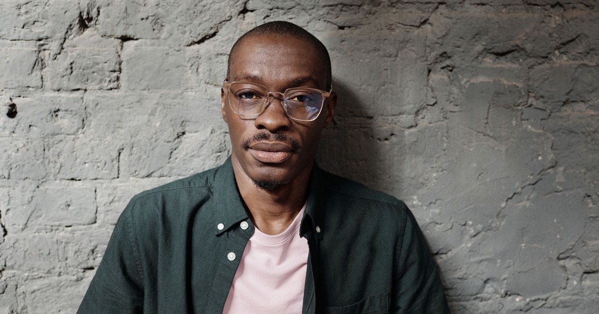 A stylish African American man wearing eyeglasses poses against a textured concrete wall.