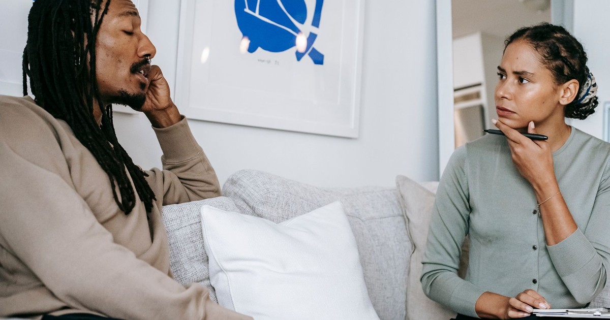 Attentive ethnic female stressed in formal wear sitting on sofa and listening to Concerned African American male client in modern psychology center