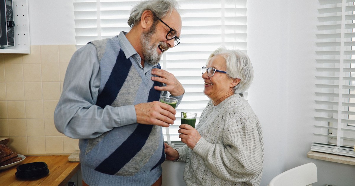 Elderly couple smiling and holding drinks in a cozy kitchen setting.