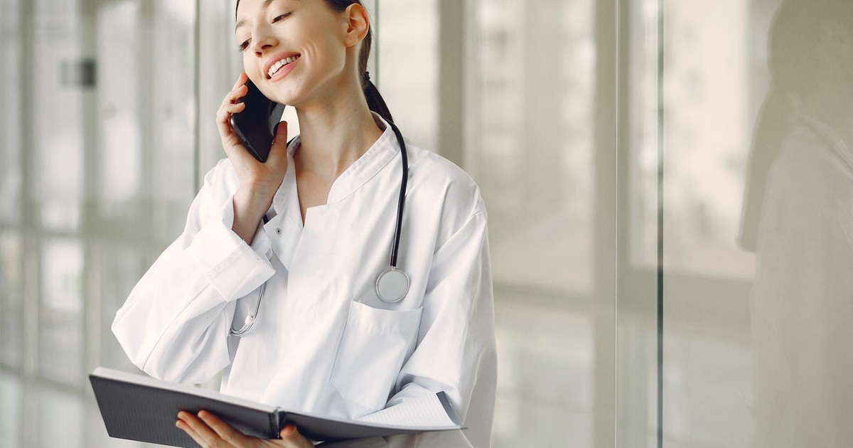 Smiling ethnic female doctor in medical uniform with stethoscope and notebook talking on smartphone while standing in glass hospital corridor