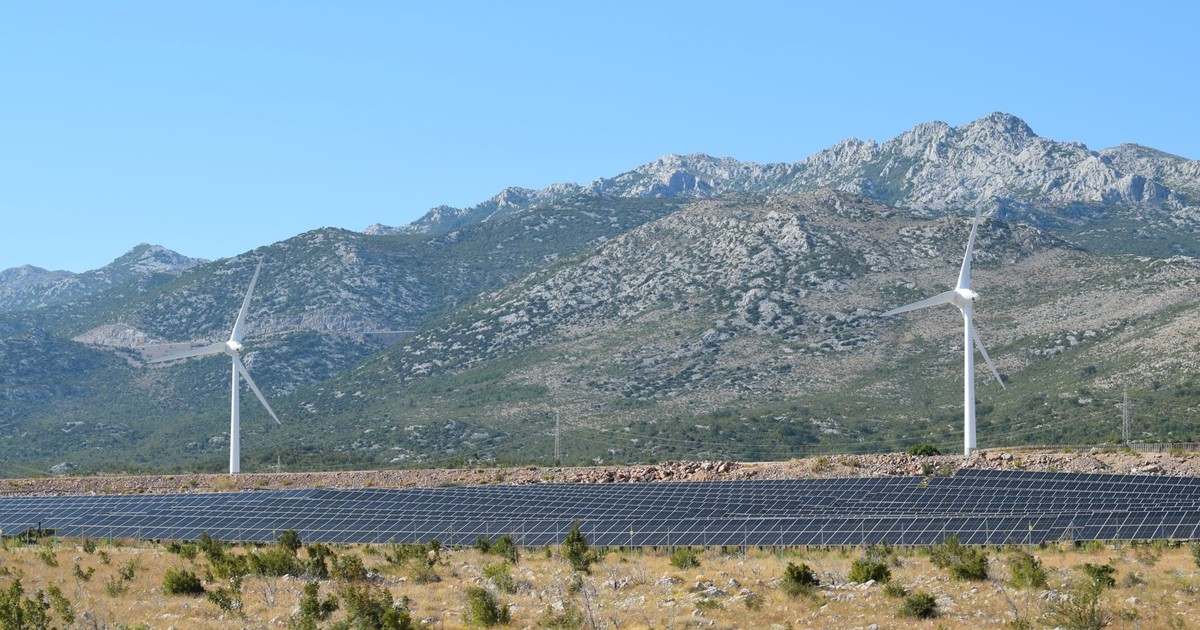A scenic view of windmills and solar panels against mountainous terrain under a clear blue sky.