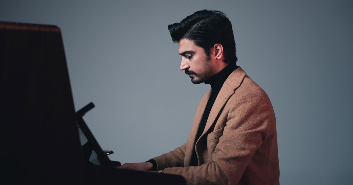 A young man in a brown coat plays piano indoors, focused and poised.