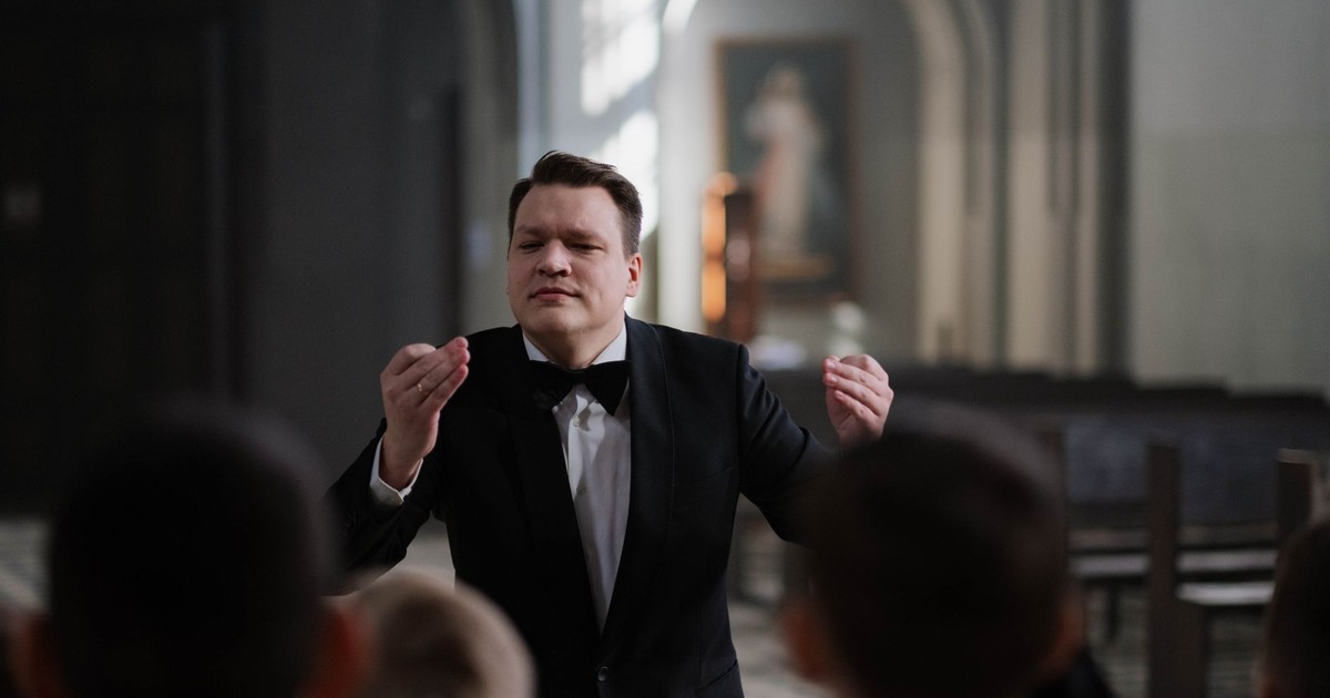 A male conductor leads a boys' choir rehearsal in a serene church setting.