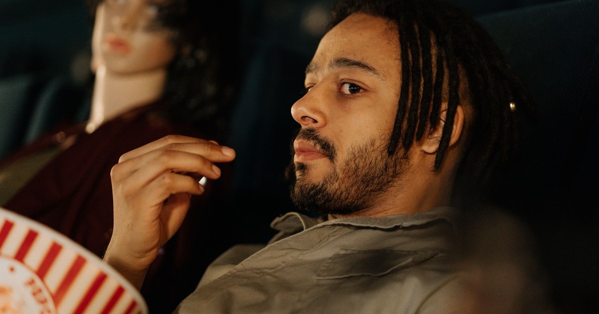 A man with dreadlocks enjoys popcorn in a cinema, sitting next to a mannequin.