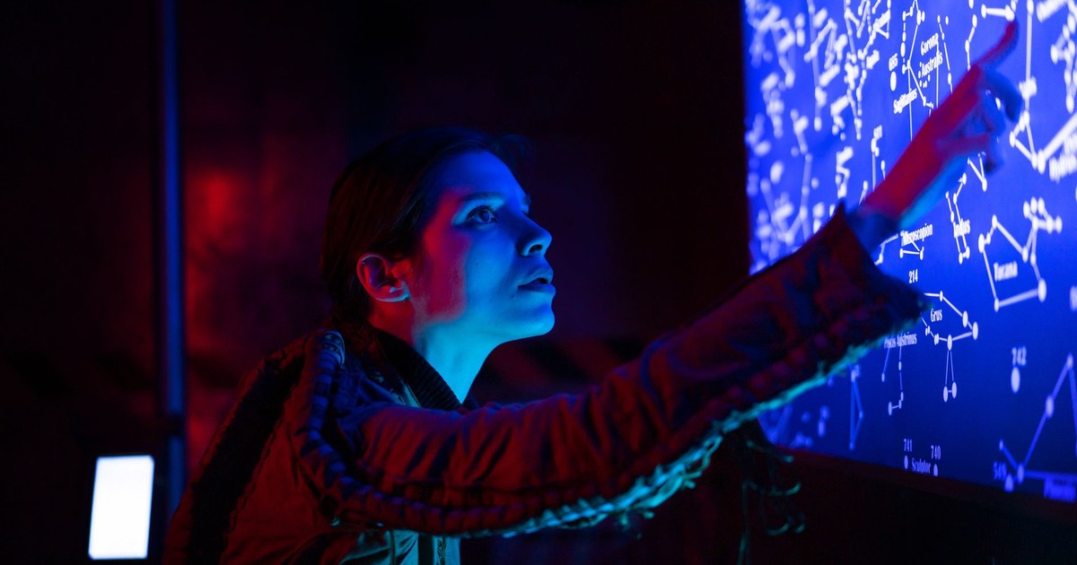 A woman studies a futuristic control panel displaying celestial charts under blue light.