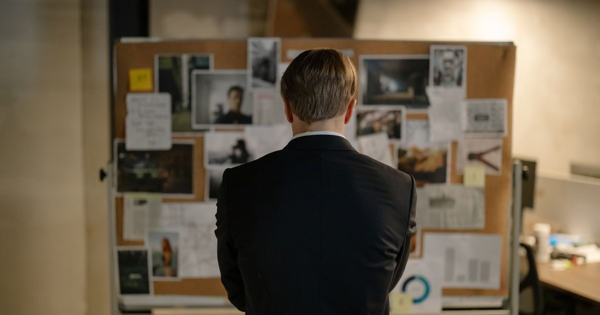 A detective in a suit examines a bulletin board with investigation materials in an office setting.