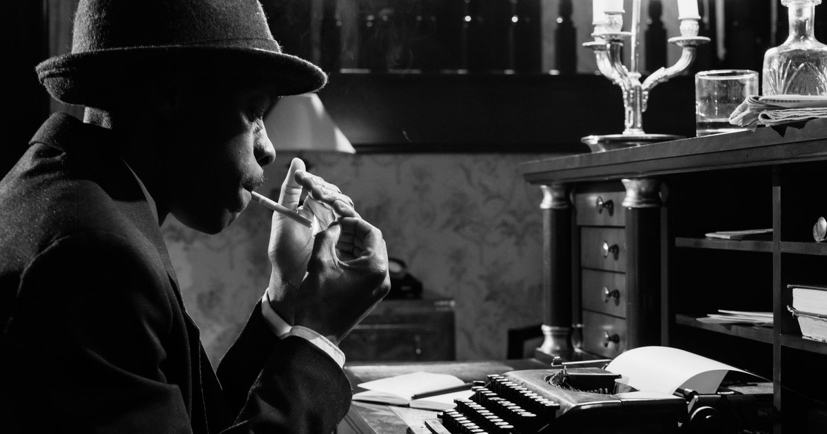 Black and white photo of a noir detective with typewriter, smoking in a vintage office setting.