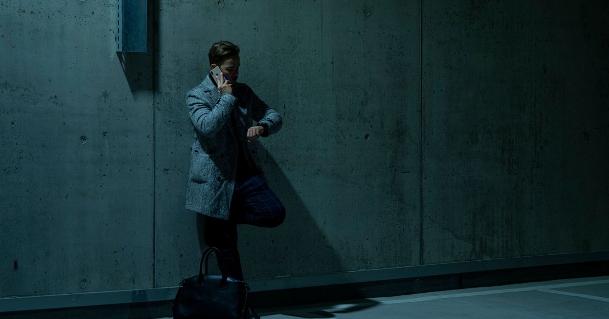 Adult man in coat on phone checks wristwatch in dim parking setting.