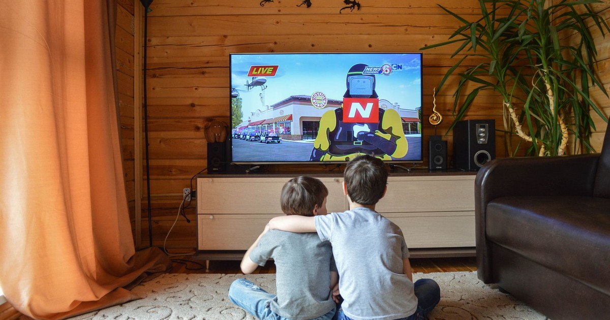 Back view of unrecognizable little children in similar casual outfits resting on floor at home and hugging while watching cartoon on TV