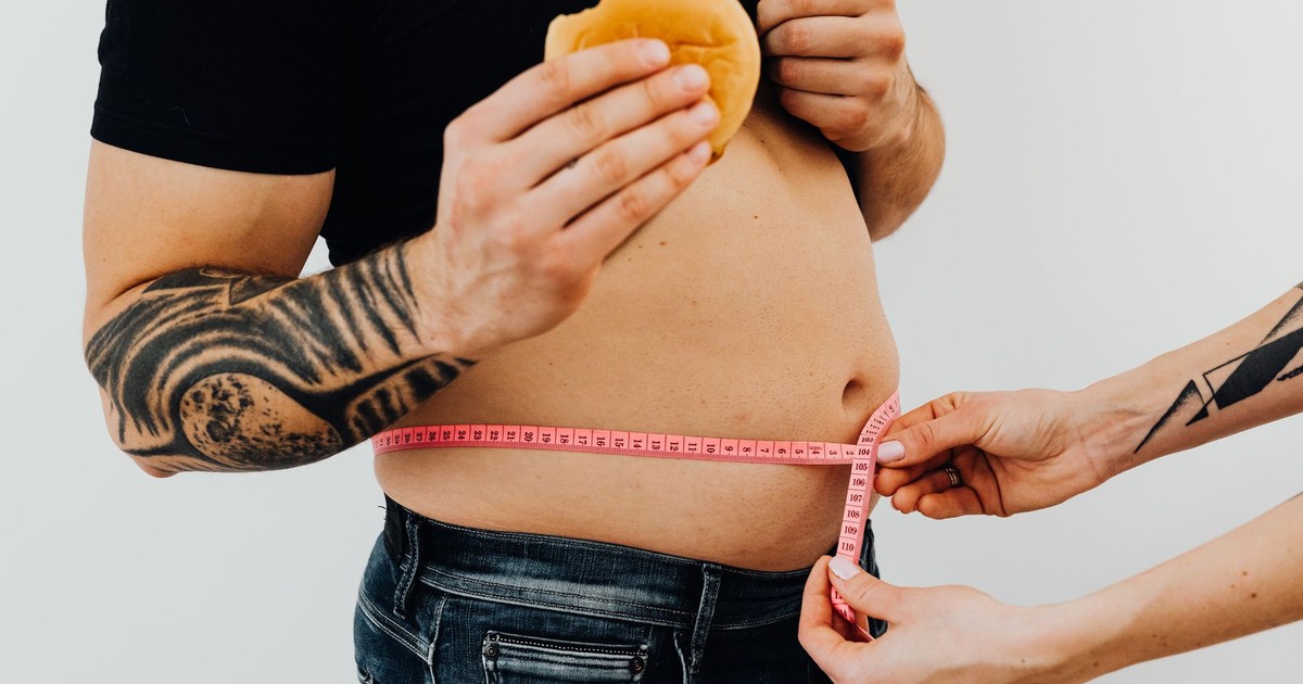 A man with tattoos holding a burger as another person measures his waistline with a tape measure.