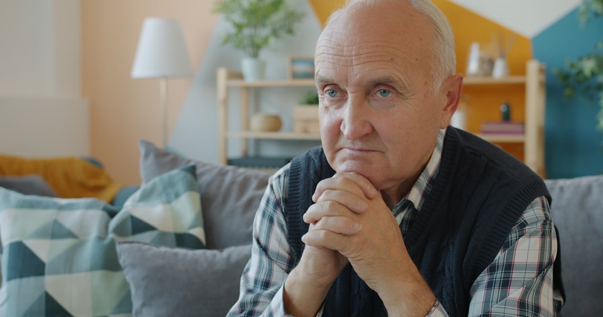 Elderly man sitting on a couch with hands clasped.