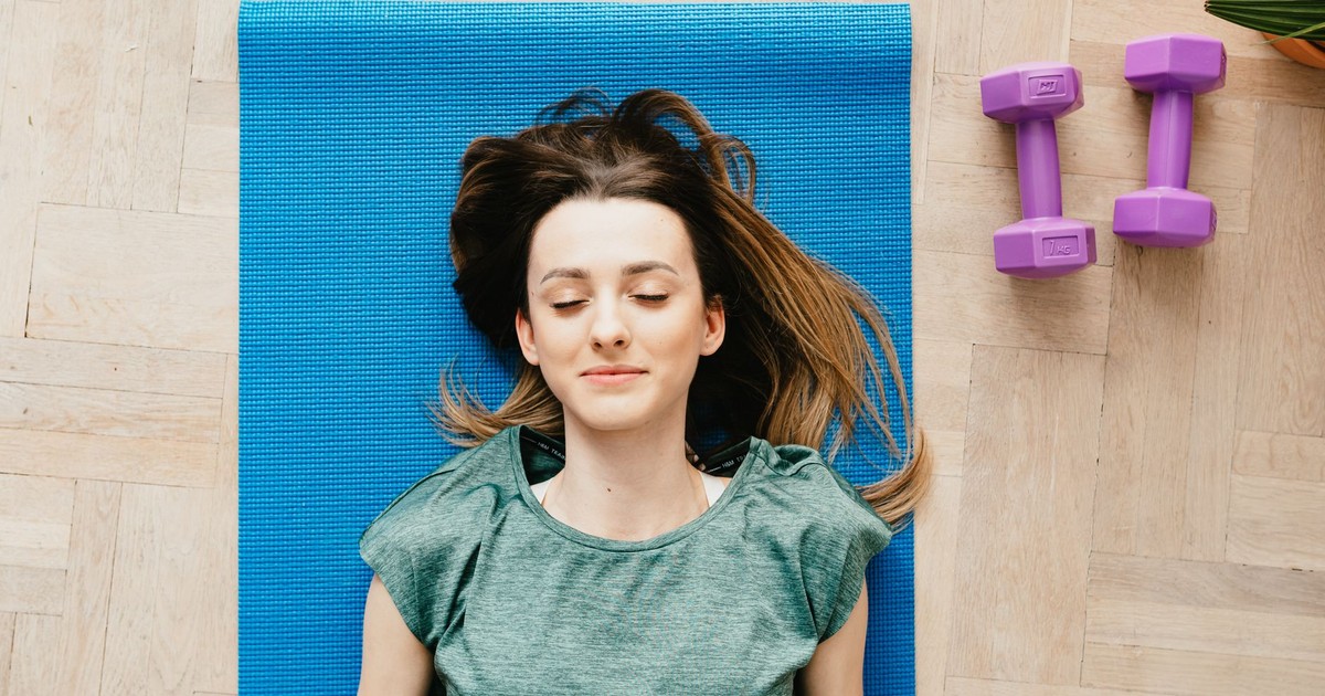 From above of content slim female in casual wear resting on blue yoga mat near dumbbells on floor at home
