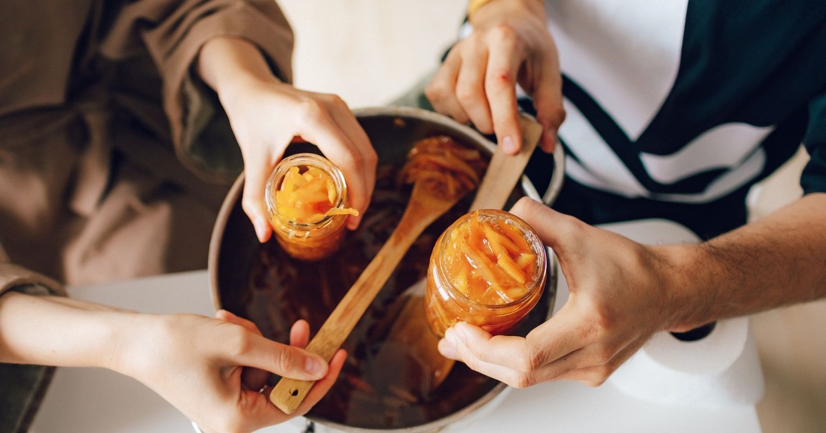 Hands making homemade orange jam, filling jars over a kitchen counter.