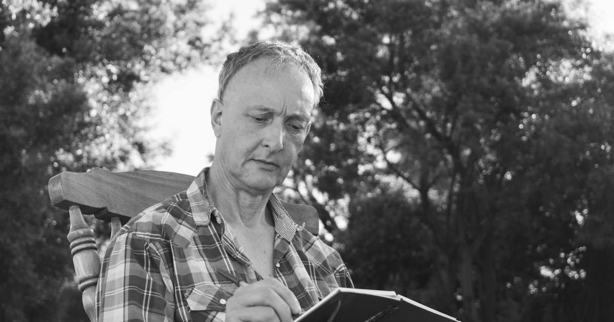An elderly man in a plaid shirt sits outdoors writing in a notebook, captured in black and white.
