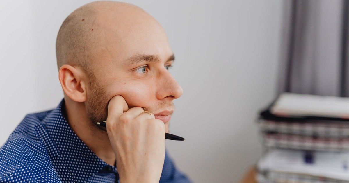 Bald man in an office thinking deeply with a pen in hand near a desk.