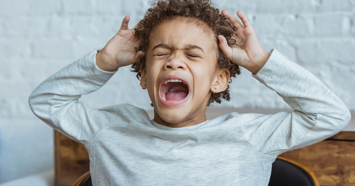 Despaired African American boy with curly hair and closed eyes screaming while standing in light room at home near wall