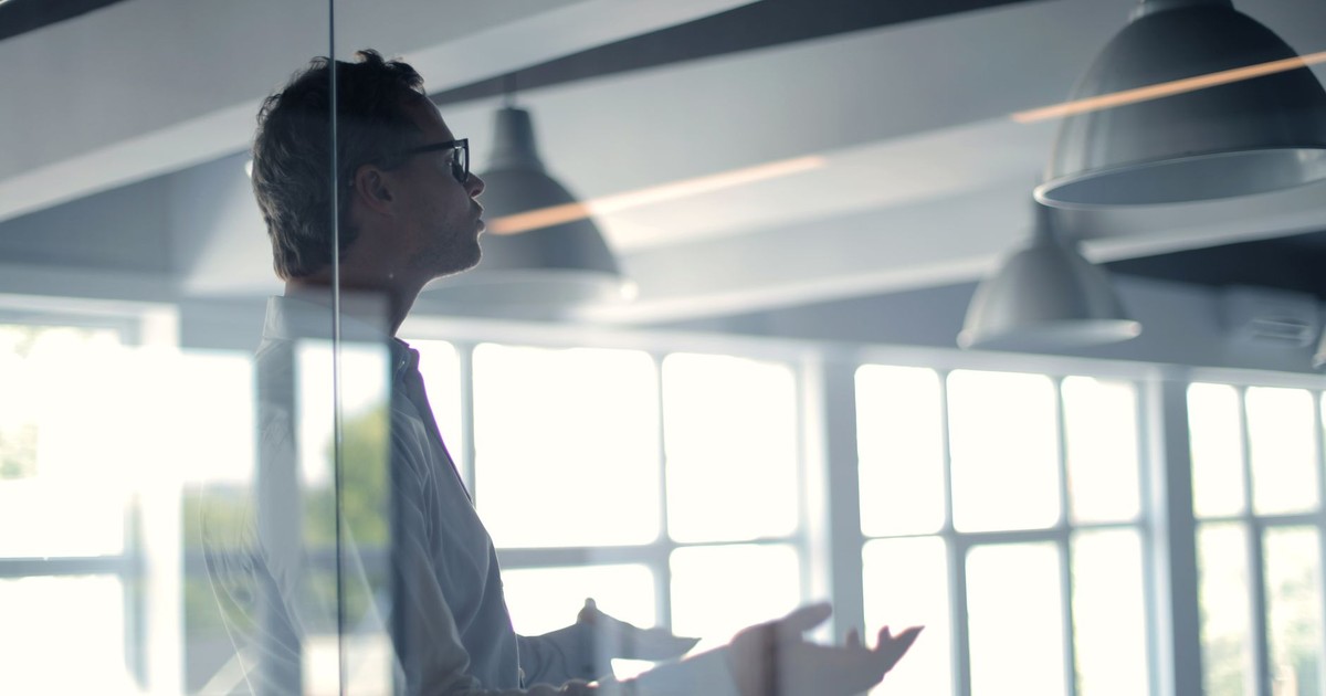 Through glass view of adult businessman in glasses and white shirt gesturing while holding presentation in spacious office