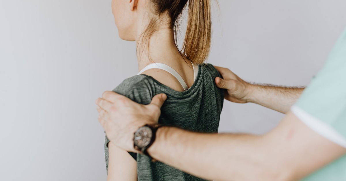 Back view of crop faceless orthopedist in uniform and wristwatch examining shoulder joints of slim female patient in clinic on white background