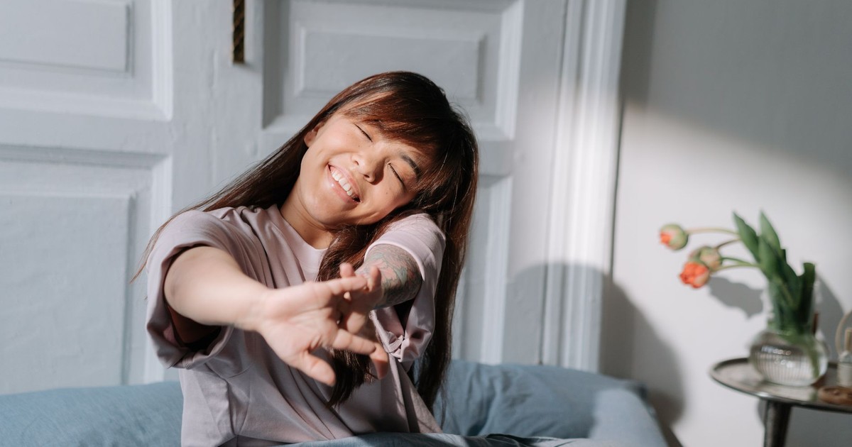 Smiling Asian woman stretching in bed, enjoying soft morning light indoors.