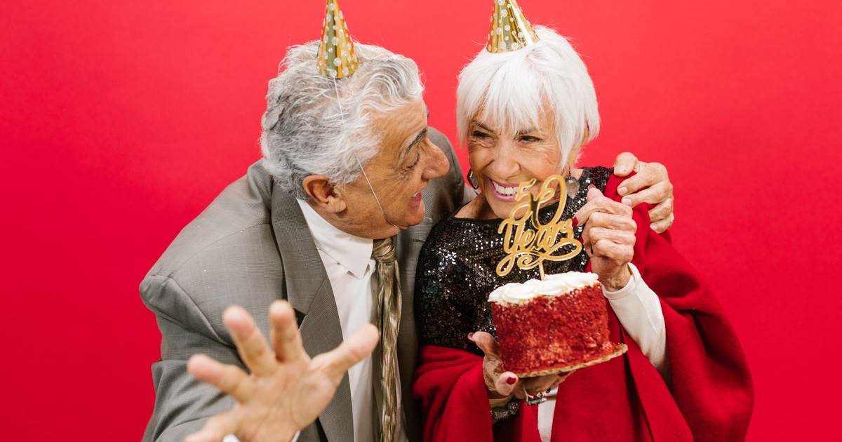 Elderly couple with party hats celebrating 50th anniversary with a cake and laughter.