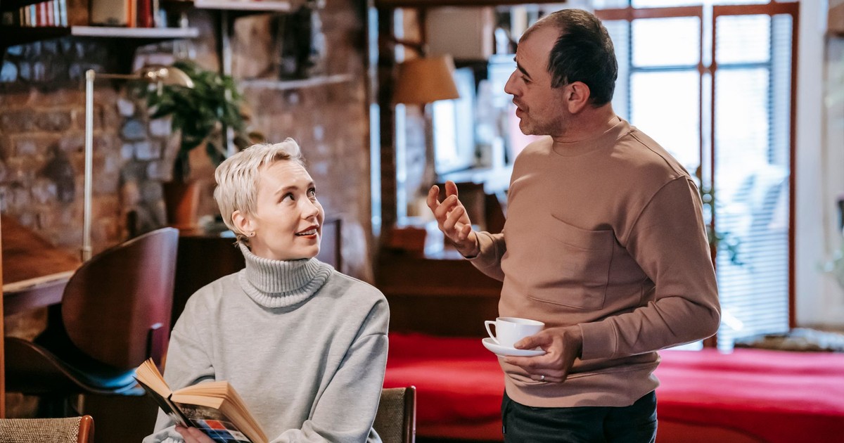 Positive diverse middle aged spouses in casual clothes drinking coffee and discussing interesting book while spending weekend together at home
