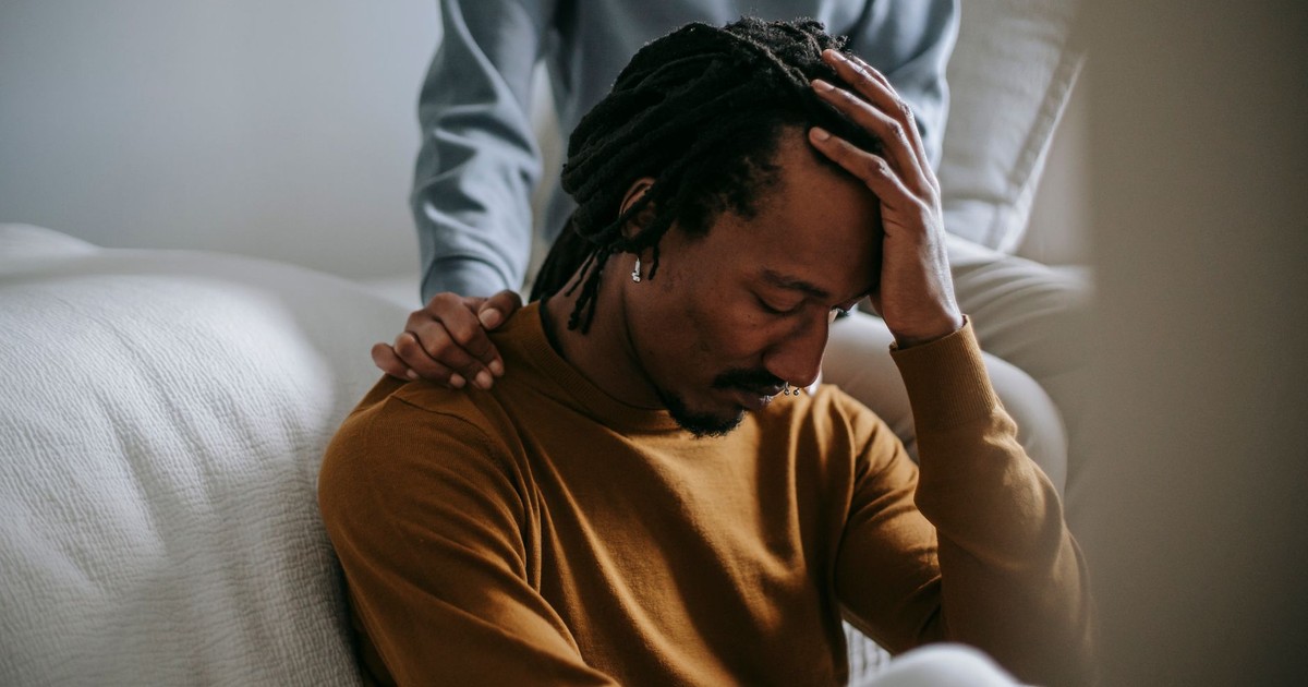 Worried depressed African American male touching head nervously and sitting with eyes closed near supporting wife in bedroom
