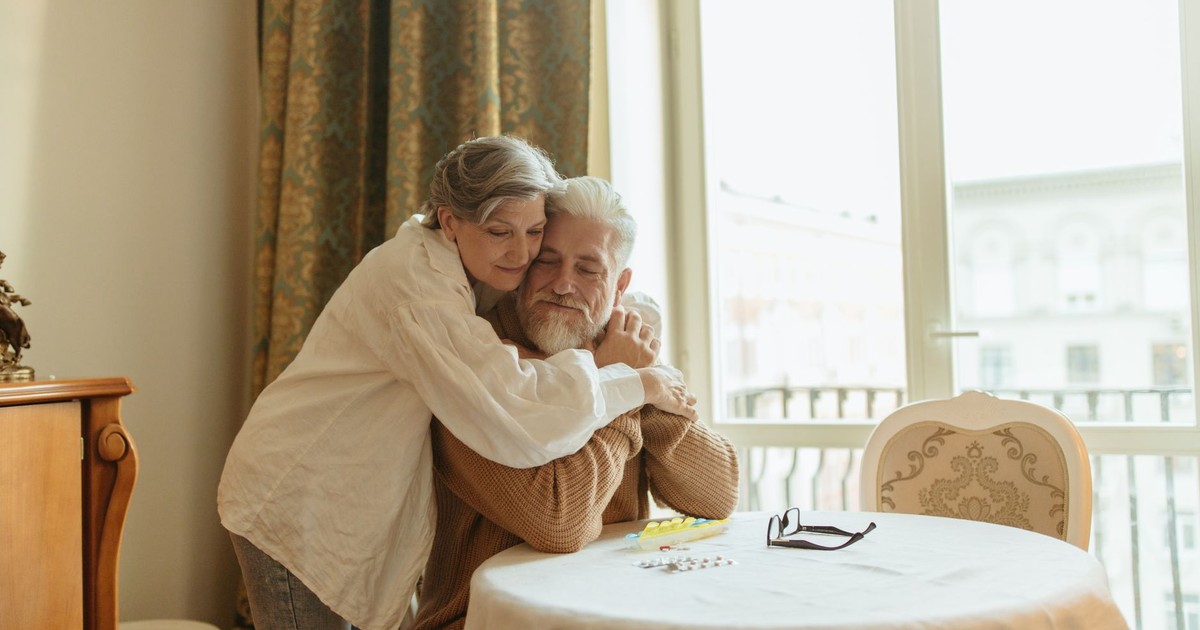 A loving elderly couple shares a warm embrace at a dining table indoors, symbolizing affection and togetherness.
