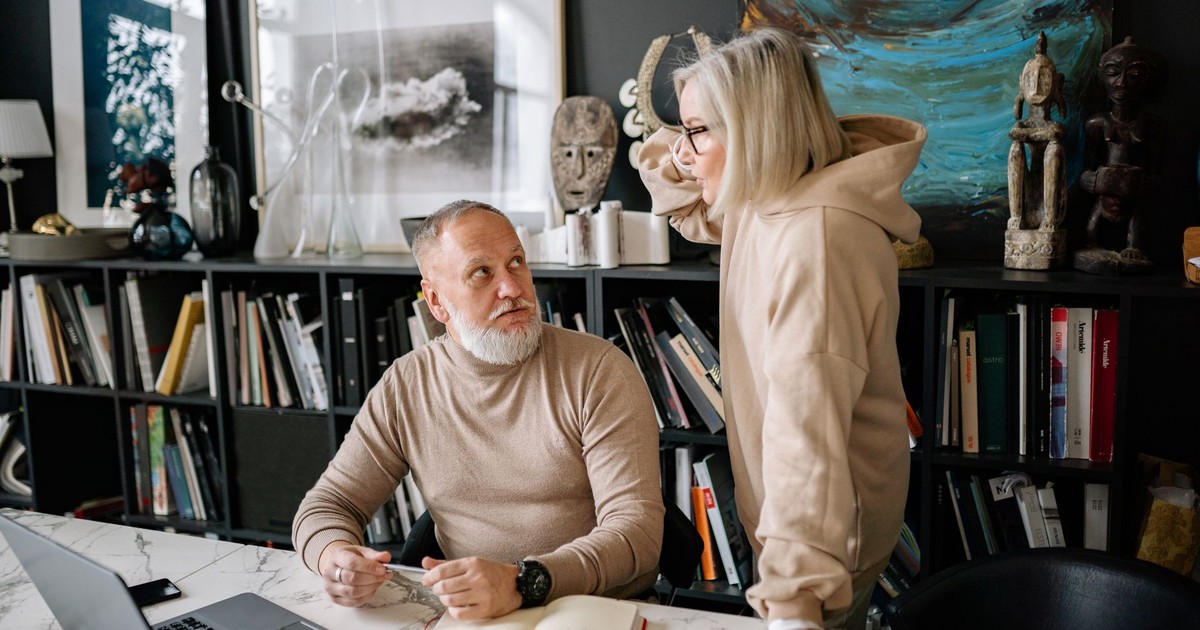 Elderly couple discussing something at a marble table with books and laptop, surrounded by art and sculptures.