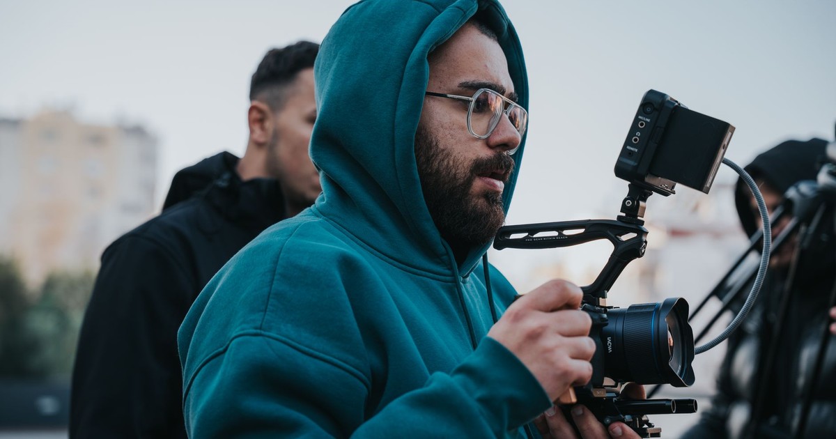 Filmmaker in blue hoodie operating a camera outdoors with a screen monitor attached.