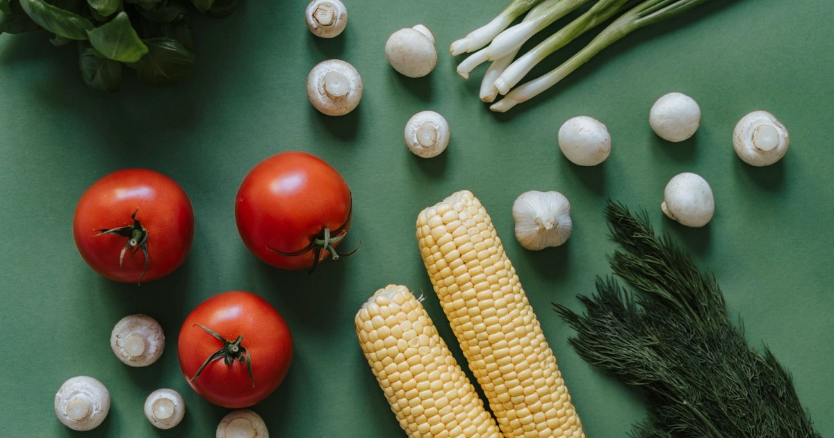A top view of fresh vegetables including tomatoes, corn, and mushrooms on a green background.