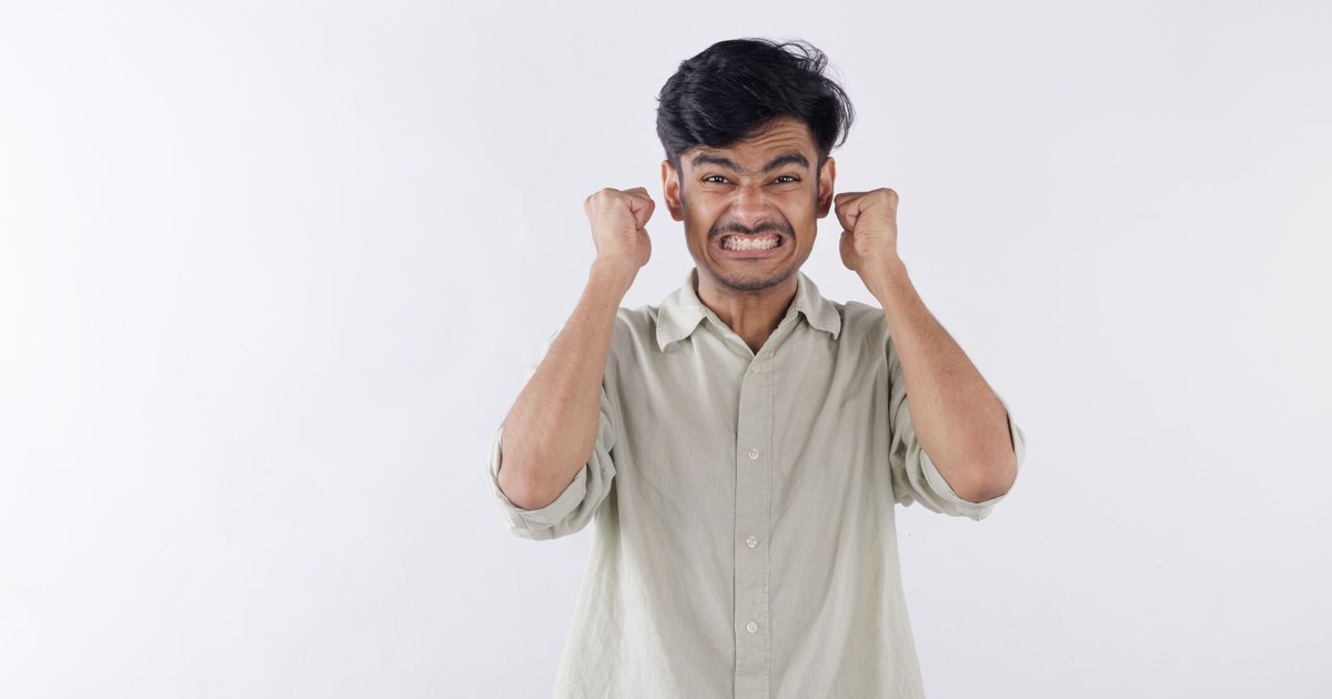 Asian man expressing frustration and stress by clenching fists against a neutral background.