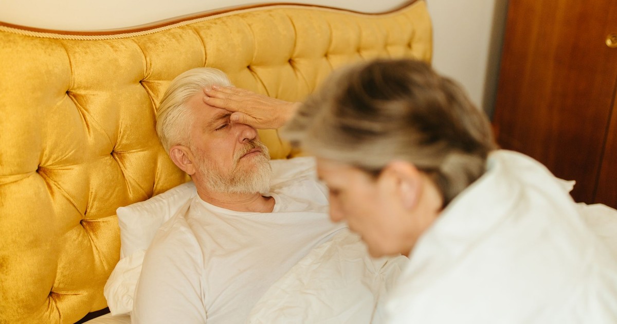 An elderly man in bed being checked for fever by a caregiver, suggesting illness or resting state.