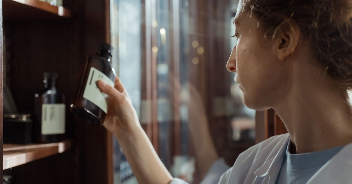 A pharmacist in a lab coat selecting a medication bottle from a pharmacy shelf indoors.