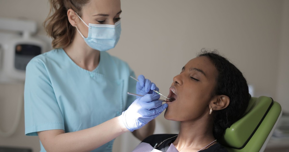Side view of young female dentist in mask and gloves wearing blue uniform holding medical tools while treating teeth of young ethnic lady in modern clinic