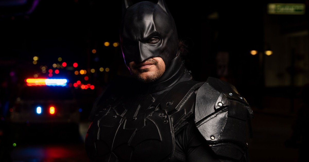 Batman cosplayer in detailed costume at a night event with police lights in the background.