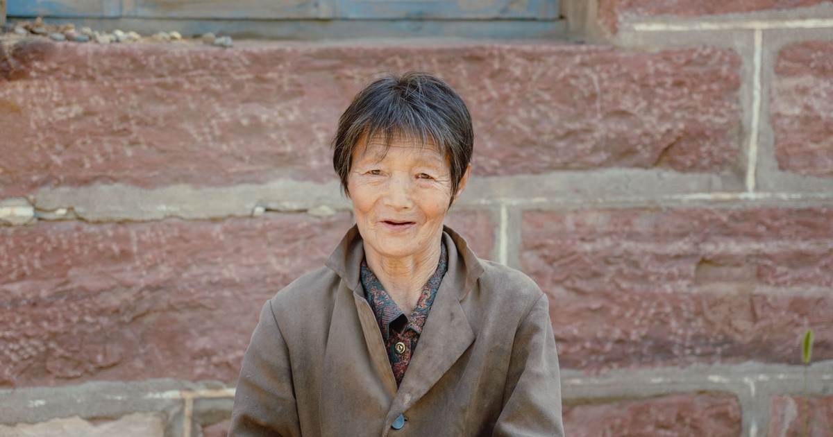 Charming portrait of an elderly Asian woman smiling against a rustic stone wall.